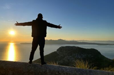 A man stands on a mountaintop with arms wide open, facing the sea and islands, preparing to practice various breathing techniques and methods.