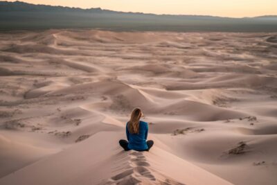 A woman sits in the middle of a desert practicing coherent breathing exercises with eyes closed, focusing on her breath, surrounded by vast, serene sand dunes.