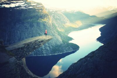 A woman practices yoga on the edge of a rock, surrounded by nature and a calm lake, embracing mindfulness and serenity.